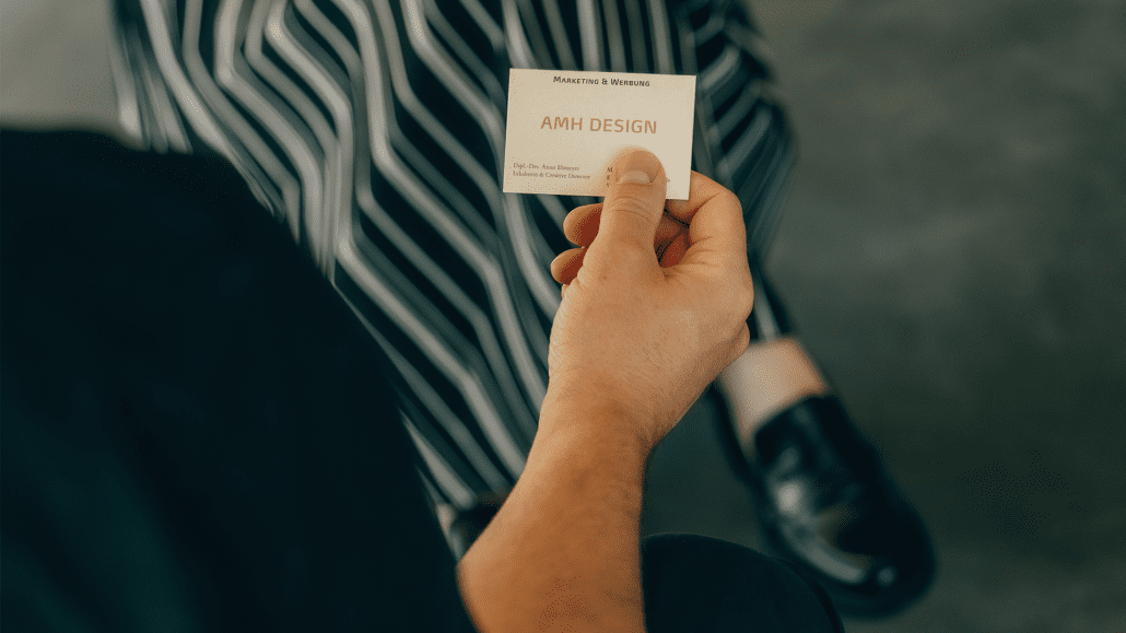 Mann hält eine Visitenkarte mit den Kontaktdaten von AMH DESIGN in der Hand, während im Hintergrund zwei Personen zu sehen sind